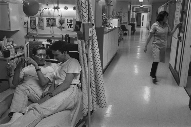 John, a man dying with AIDS, lies on a hospital bed with his partner, both dressed in t-shirts and jeans. A nurse walks by in the corridor. Greeting cards are pinned on the wall behind the bed.