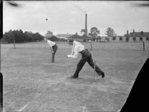 On a cricket pitch, a man with his left arm in plaster bowls overarm with his right arm.