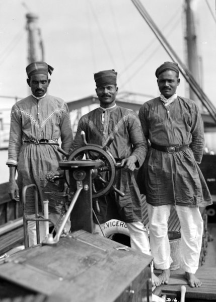 Three Lascar sailors, two barefeet, stand behind a ship's helm
